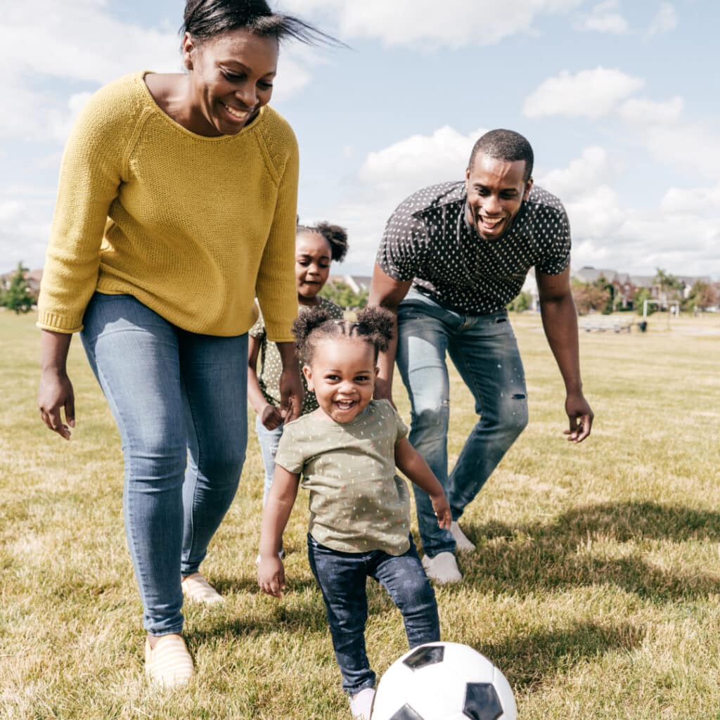 Family of four playing soccer in a sunny park; toddler centered, about to kick the ball, as mom and dad smile nearby on the grass with an older child behind them.