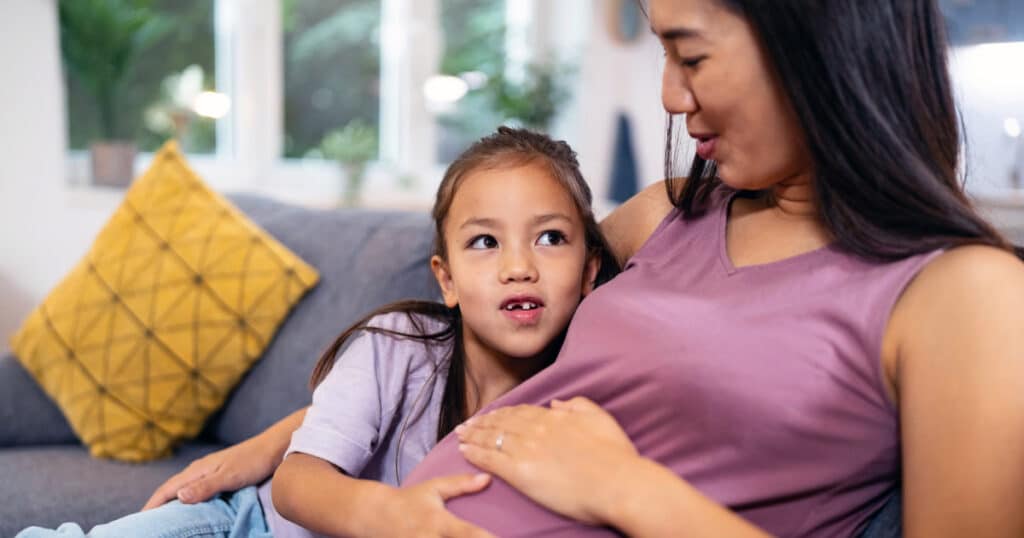 A pregnant woman sits on a couch while a young girl affectionately rests her head on her belly and looks at her, sharing a tender moment.