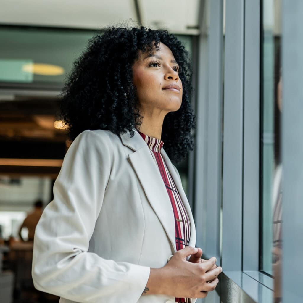 Woman with curly hair in a cream blazer gazing out a glass window in a modern office setting. (informative)