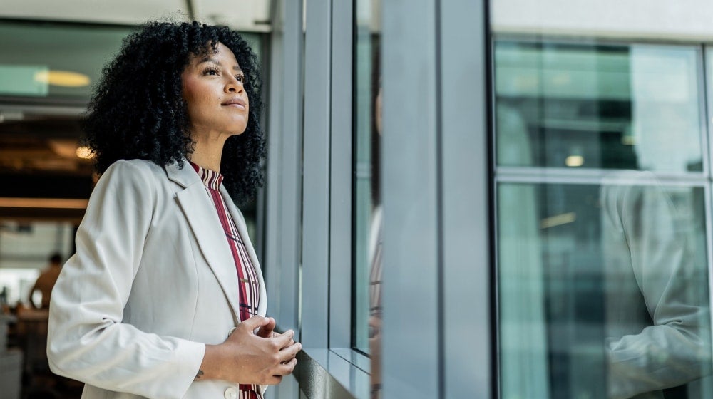 Thoughtful professional woman standing in a modern office, reflecting during a moment of workplace change and uncertainty.
