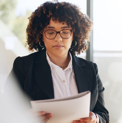Professional woman with curly hair and glasses reads a document in a blazer by a window, focused on the papers.