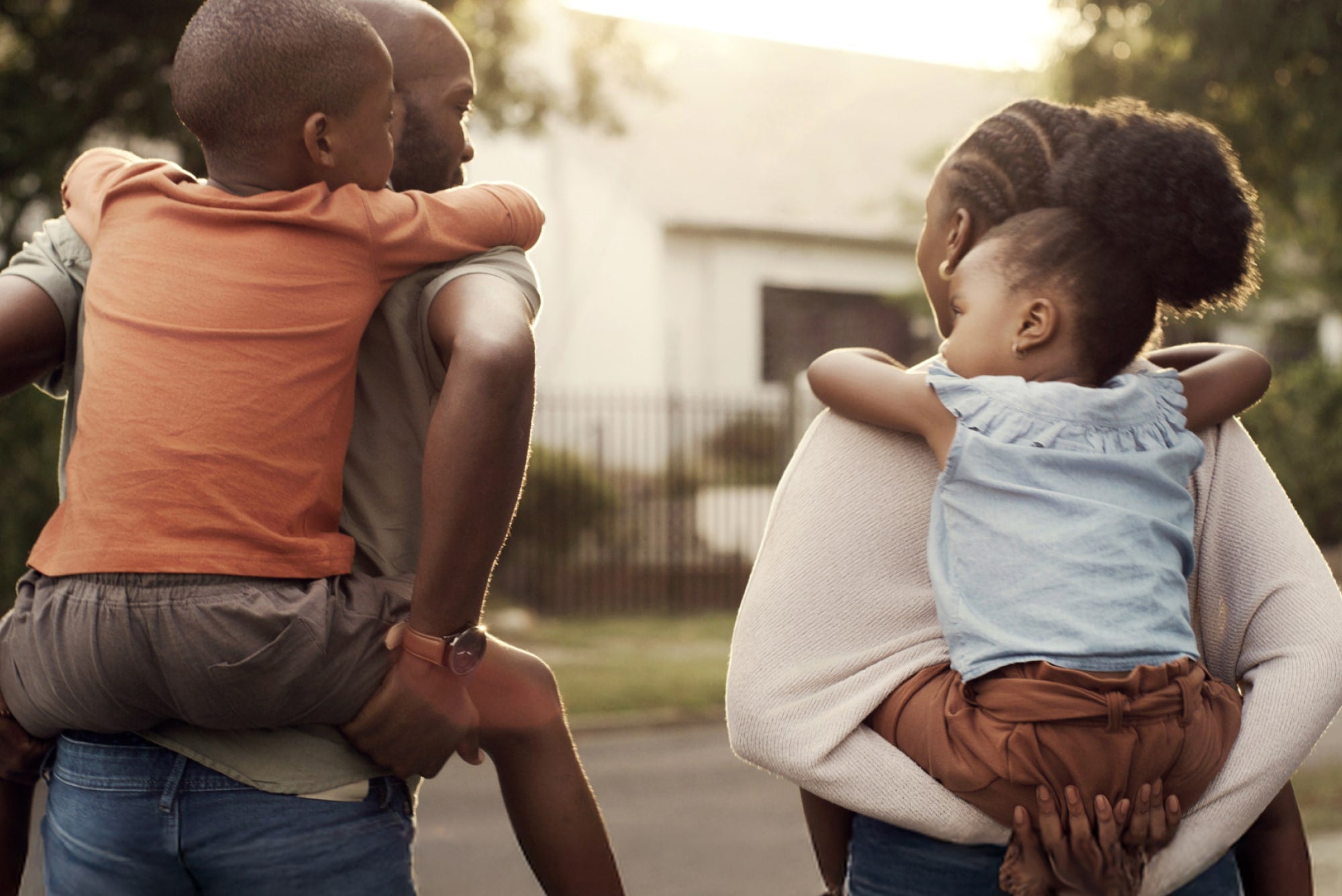 Two parents carry their children on their shoulders outdoors at sunset in a residential neighborhood.