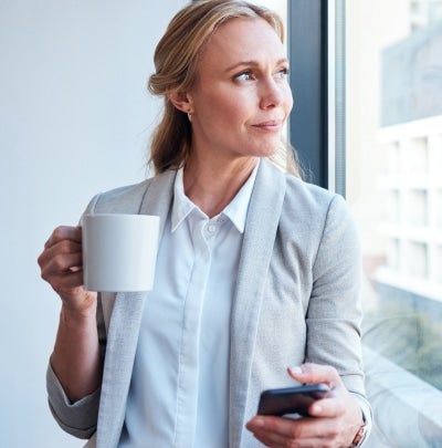 Mature professional woman looking out an office window while holding a smartphone, reflecting on future financial goals and career planning.