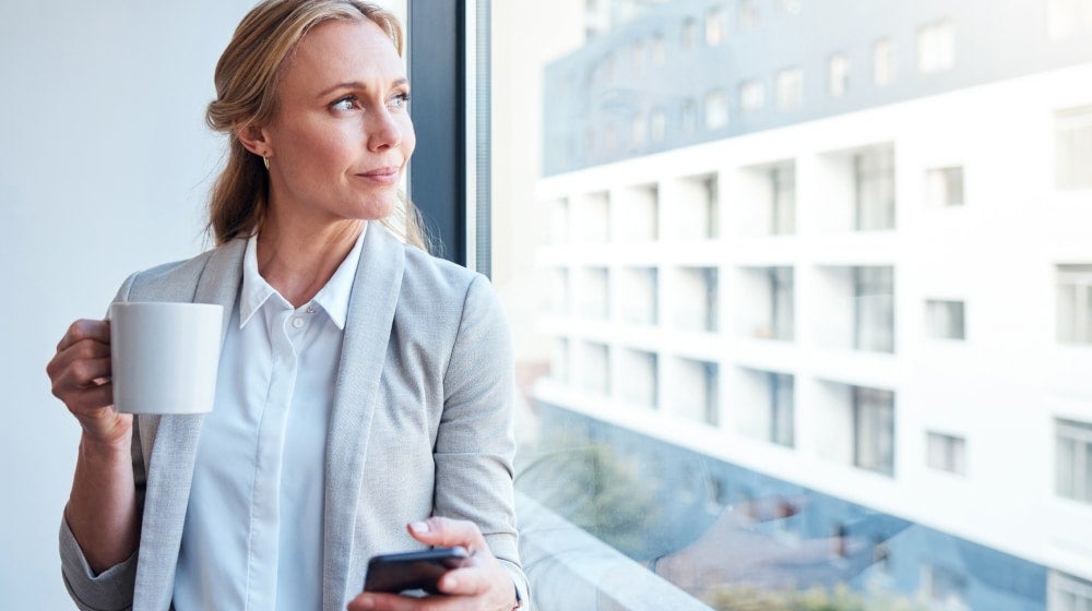 Mature professional woman looking out an office window while holding a smartphone, reflecting on future financial goals and career planning.