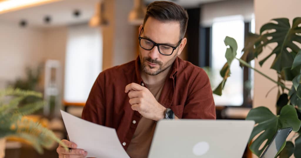 Man with glasses reviews a document at a desk in a modern office with a laptop nearby and plants in the background, looking thoughtful.