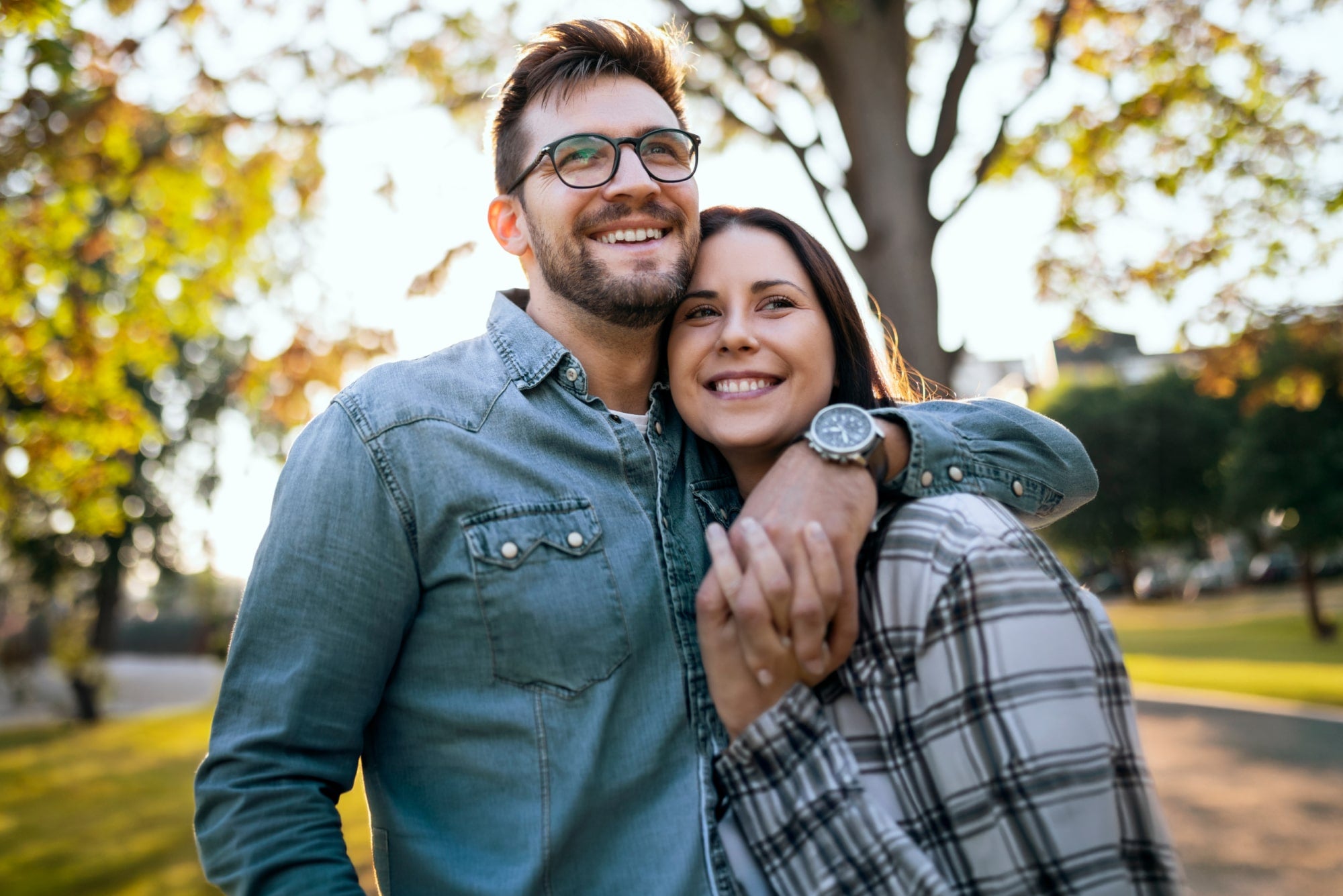 Couple smiling and hugging in a sunlit park with autumn leaves in the background.