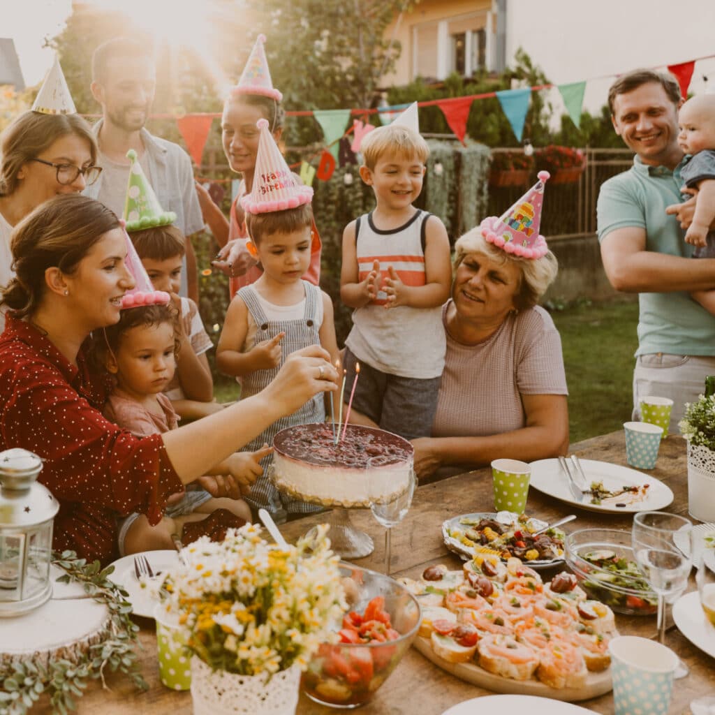 Family and friends at an outdoor birthday party, kids wearing party hats around a cake with candles on a wooden table, with snacks and colorful cups nearby.