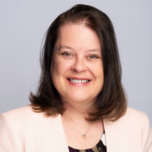 Head-and-shoulders portrait of a smiling woman with brown hair in a light blazer against a neutral background.