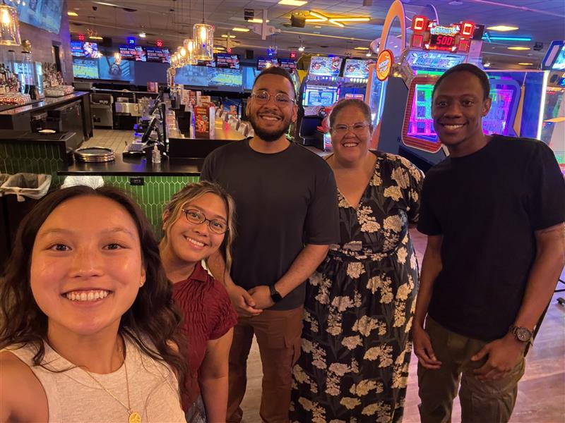 Five friends and family members pose for a group selfie in a bowling alley, with neon arcade games and bowling lanes in the background.