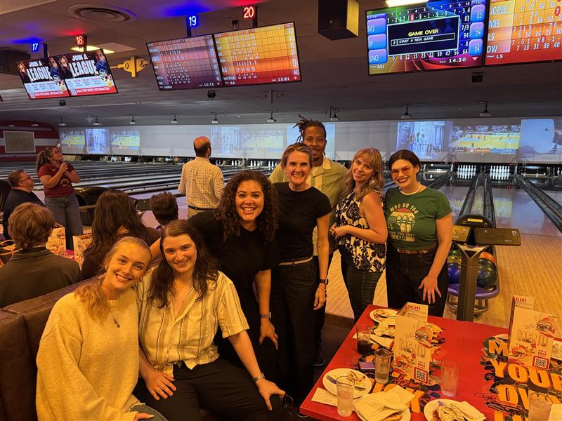 Group of friends posing and smiling near bowling lanes at a social night, with a table of food and drinks in the foreground.