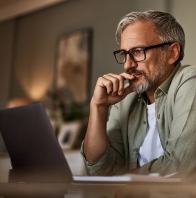 Older man with gray hair and glasses sits at a desk, thinking while looking at a laptop in a home office.