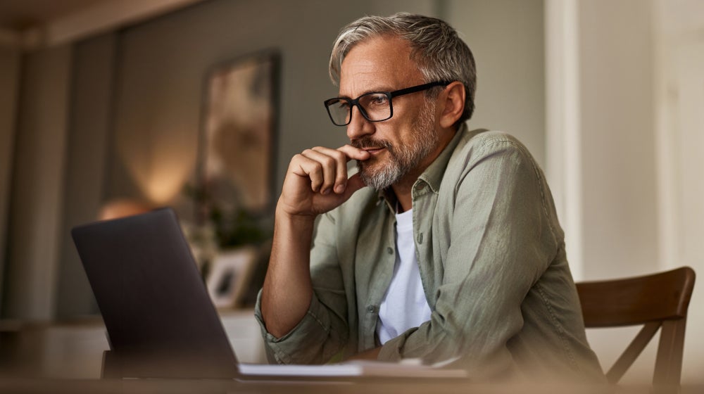 Man with gray hair and glasses thinking while using a laptop at home.