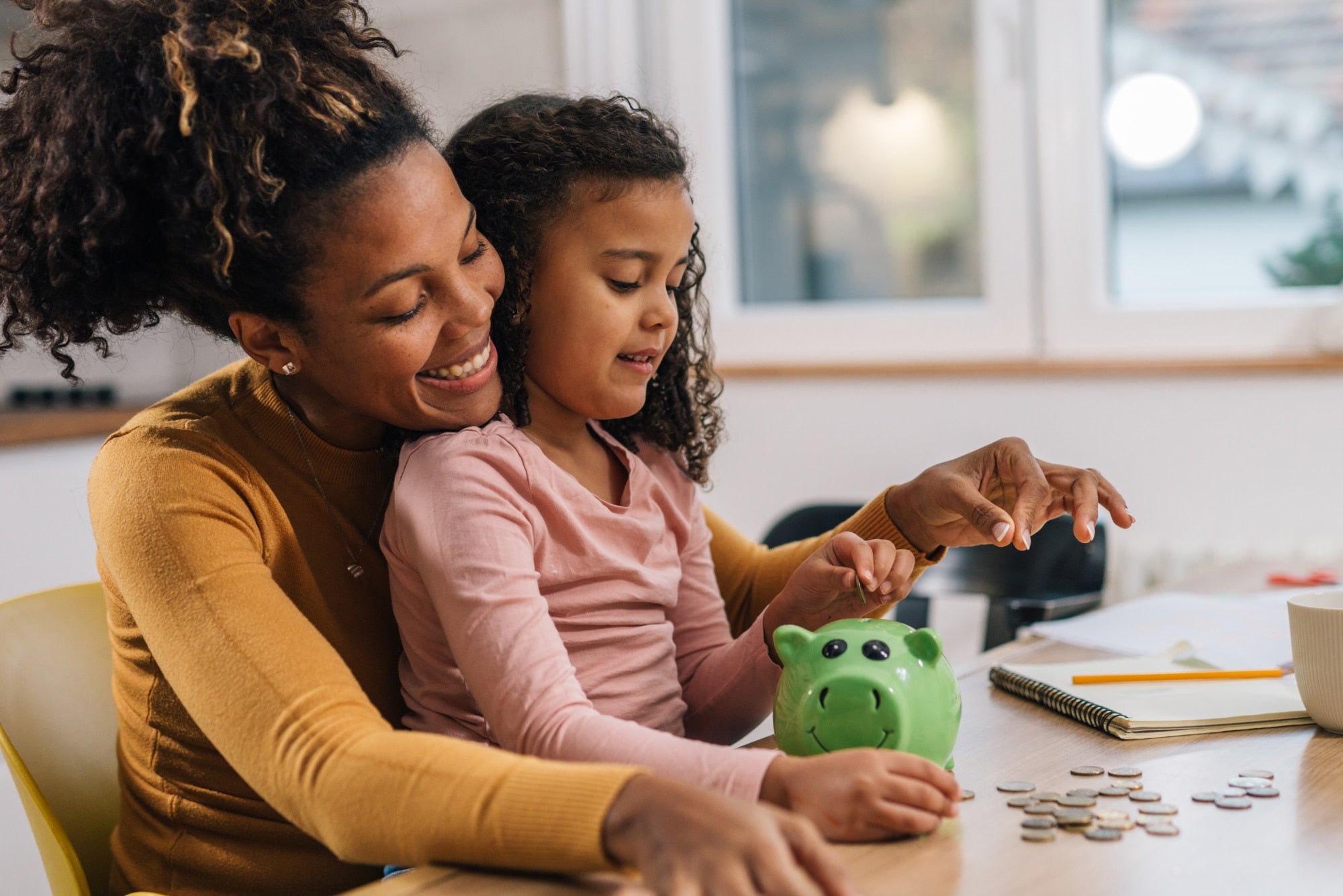 Woman and child counting coins at a table with a green piggy bank nearby.