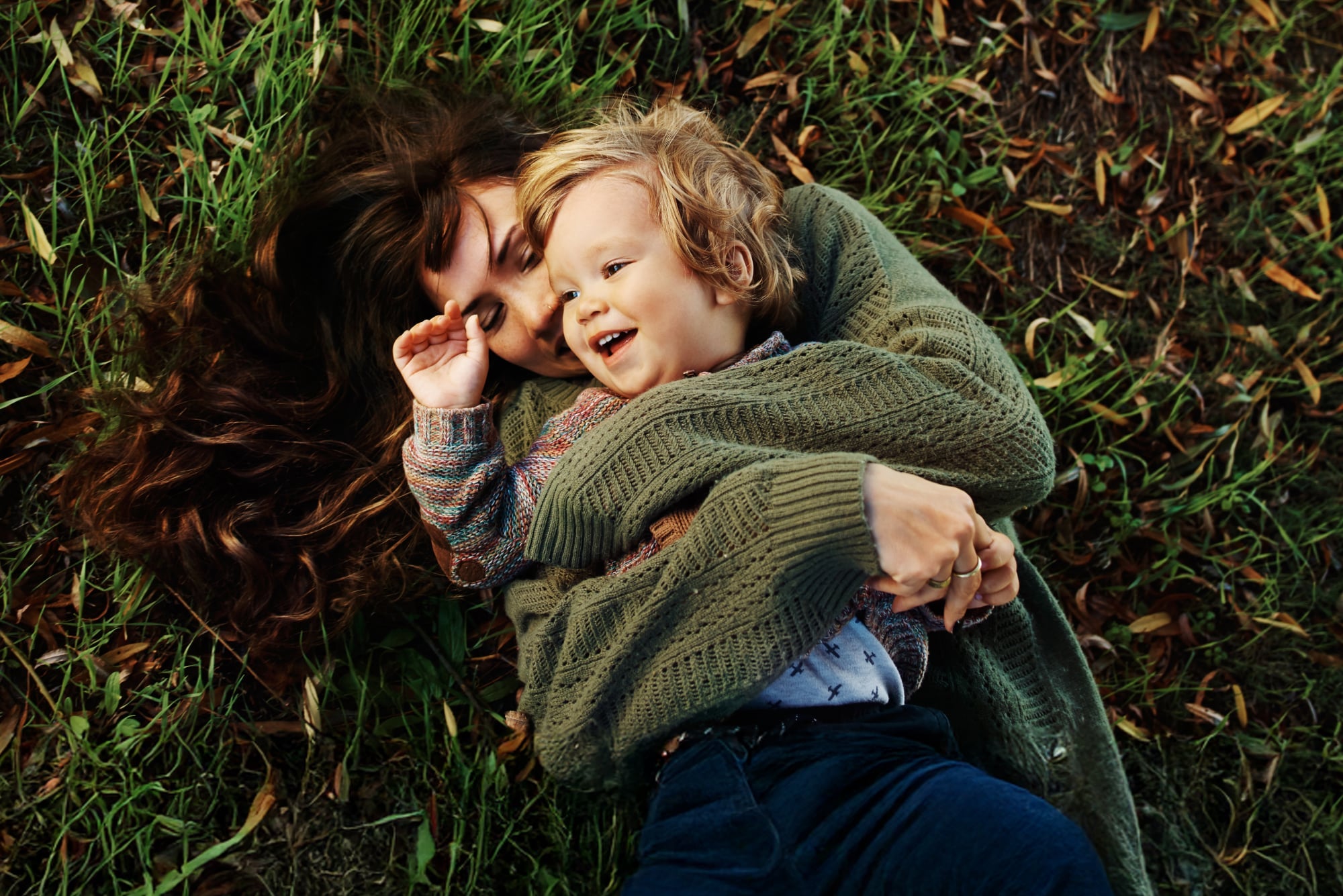 A mother and young son laying together in the grass in an embrace.